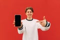 Positive guy in a white sweatshirt stands on a background of a red wall, looks at the camera and shows his index finger on a Royalty Free Stock Photo