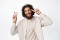 Positive handsome guy smiling, showing v-sign peace gesture and laughing, standing happy against white background Royalty Free Stock Photo