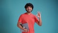 Positive handsome indian or arabian curly-haired guy, student, freelancer, holds an laptop, points OKAY gesture Royalty Free Stock Photo