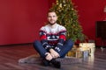 Positive handsome man sits near Christmas tree and wrapped gift box present from santa claus Royalty Free Stock Photo