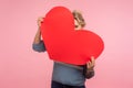 Positive happy woman with curly hair hiding face behind big red heart and looking at camera with curious prying eyes