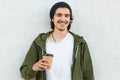 Positive hipster in stylish black headgear, white t shirt and anorak, holds paper cup of coffee, enjoys spare time, isolated over
