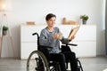 Positive impaired teenager in wheelchair drinking tea and reading interesting book at home