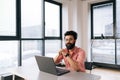 Positive Indian businessman in casual clothes sitting at desk with laptop smiling looking at camera in light coworking Royalty Free Stock Photo