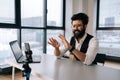 Positive Indian male vlogger in glasses broadcasting from office, using smartphone and tripod, sitting at table with Royalty Free Stock Photo