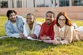 Positive international millennial students exchange, rest outdoor, lie on grass in campus, enjoy break