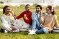 Positive international millennial students study drink coffee, relax, sit on grass together in campus Royalty Free Stock Photo