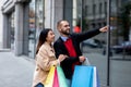 Positive interracial couple walking along street with paper bags, pointing at new store window, shopping together Royalty Free Stock Photo