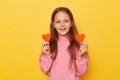 Positive joyful brown haired little girl wearing pink sweatshirt isolated over yellow background showing two little red hearts