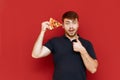 Positive man with beard stands on red background with piece of pizza in hand, looks into camera, smiles and shows thumb up. Hungry Royalty Free Stock Photo