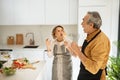 Positive married senior couple having fun while cooking in kitchen interior, singing and dancing, using untensils as mic