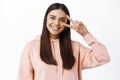 Positive millennial girl with candid smile, showing v-sign on eye with happy face, standing against white background