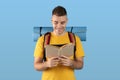 Positive millennial hipster with camping gear reading dictionary or guidebook over blue studio background