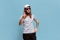 Positive mood. Portrait of young smiling man with long hair posing in cap and sunglasses against blue studio background Royalty Free Stock Photo