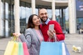 Positive multiracial couple with bright gift bags and cellphone placing online order near shopping mall Royalty Free Stock Photo