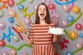 Positive overjoyed woman with brown hair wearing striped dress holding cake and smart phone in hands, screaming with excitement,