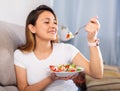 Peruvian housewoman eating vegetable salad on sofa at home