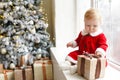 Positive photo of cute beautiful little girl in Santa`s dress playing christmas gift box on the windowsill Royalty Free Stock Photo