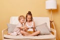 Positive playful little girls sisters sitting on couch using smartphone together posing against beige wall watching online video Royalty Free Stock Photo