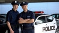 Positive police mates in hats smiling into camera against squad car, work