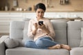 Positive pretty Indian woman resting on home sofa with smartphone