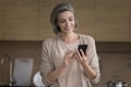 Positive grey haired woman holding smartphone in home kitchen Royalty Free Stock Photo