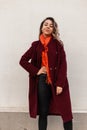 Positive pretty young woman fashion model with vintage orange scarf in stylish burgundy coat posing near vintage wall. Fashionable Royalty Free Stock Photo