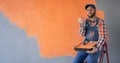 Positive professional male painter with roller pointing at blank space on orange wall sitting on ladder and looking at
