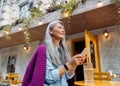 Positive senior Asian lady holds smartphone sitting at small yellow table outdoors