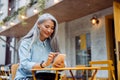 Positive senior Asian woman surfs internet with phone sitting at yellow table