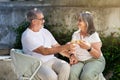 Positive senior caucasian man and lady cheers glasses of wine sit on chairs in home garden