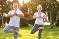 Positive senior couple performing yoga on green lawn in park during sunrise