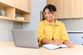 Positive smart young mixed-race female student smiling sitting at the table with laptop in the kitchen, studying from Royalty Free Stock Photo