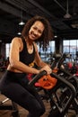 Positive smiling african american woman, exercising on an exercise bike in the gym, dark background, cardio training on Royalty Free Stock Photo