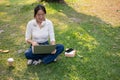 A positive, smiling Asian woman is using her laptop while sitting on the grass in a green park Royalty Free Stock Photo