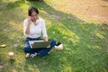 A positive, smiling Asian woman is using her laptop while sitting on the grass in a green park Royalty Free Stock Photo