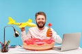 Positive smiling bearded man sitting at workplace with lifebuoy holding bottle with lemonade and toy plane, travel agent Royalty Free Stock Photo