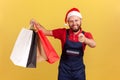 Positive smiling delivery man in santa claus hat holding paper bags and pointing finger at you, asking to use delivery service