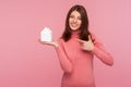 Positive smiling woman with brown hair in pink sweater pointing finger at paper toy house in her hand, apartments rent and sale,