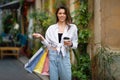 Positive stylish young millennial caucasian lady tourist hold many packs of purchases and coffee cup, enjoy shopping