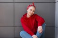 Positive stylish young woman with hairstyle in a red bandana in a fashionable red sweatshirt in vintage jeans sits and smiles