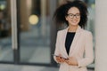 Positive successful woman entrepreneur with Afro hair holds digital tablet, stands outdoor near office building, wears formal Royalty Free Stock Photo