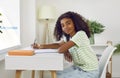 Positive teen african american girl is doing her homework sitting at desk at home lookinh at camera. Royalty Free Stock Photo