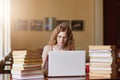 Positive tender sweet girl with curly hair working on her laptop being in reading hall with huge bunch of books on desk, focused