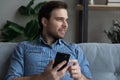 Positive thoughtful cellphone user sitting on sofa at home