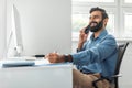 Positive thoughtful indian businessman talking on cellphone and taking notes while working on computer in office Royalty Free Stock Photo