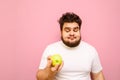 Positive totsun in a white t-shirt with curly hair and beard stands with an apple in his hand on a pink background and looks at Royalty Free Stock Photo