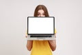 Positive unknown female in yellow T-shirt hiding half of face behind laptop with white empty display Royalty Free Stock Photo