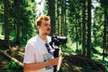 Positive video maker tourist with a stabilizer and a camera in his hands stands in the woods and looks away with a smile on his