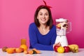 Positive woman with cheerful facial expression, being in high spirit, mixes fruit in blender for making smoothie, dressed in blue Royalty Free Stock Photo